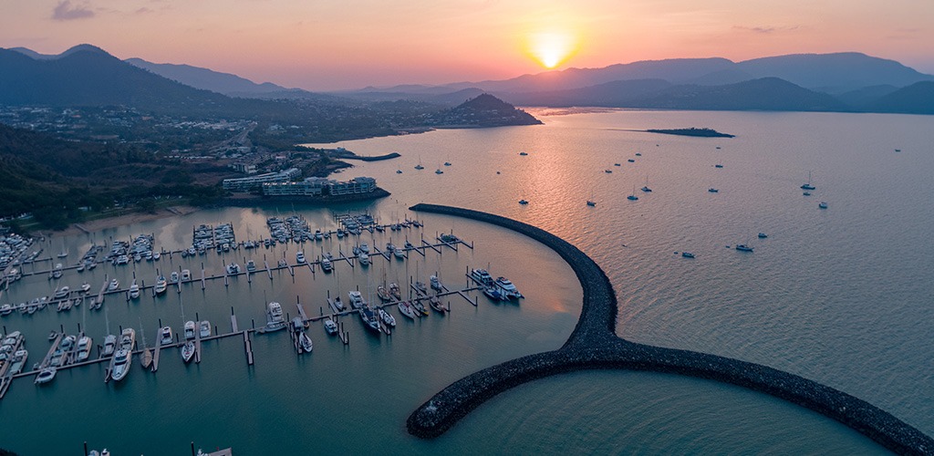 Airlie Beach marina in the Whitsundays at sunset, with boats anchored against a backdrop of coastal mountains.